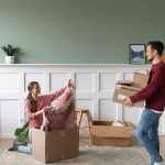 Couple unpacking boxes in new home, woman holding blanket, man carrying boxes, green wall and plant in background